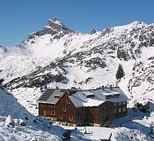 Schrägaufnahme einer T-förmigen Berghütte, der Freiburger Hütte, vor schneebedeckten Bergen