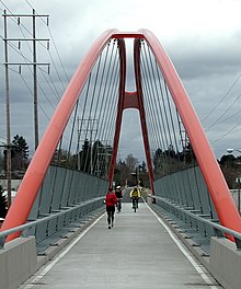 Two bicyclists and a jogger cross a long pedestrian bridge. The bright orange supporting arches of the bridge meet in the air about 30 feet (9.1 meters) above the bridge deck. Beyond the far end of the bridge is a paved bike path that continues into a woods in the distance.