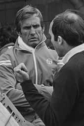 Photograph of Carlos Reutemann in the background in conversation with Frank Williams, visible in the foreground with his head turned away from the camera.