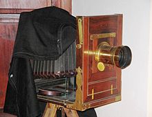 Farbfotografie einer Balgenkamera mit schwarzem Tuch über Sucher und teilweise den Balgen. Der vordere Teil mit Objektiv besteht aus Holz und goldenen Scharnieren. Die Kamera steht auf einem Holzstativ.