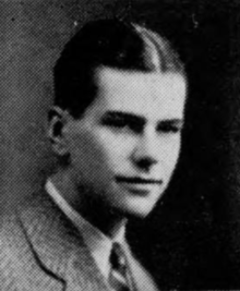 A black-and-white photo of a man in a gray suit with a tie at bust length, with hair parted in the middle and slicked, with his body oriented towards the left and his head turned halfway towards the camera