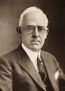 A black-and-white photo of a middle-aged Anglo man with white, receding hair, round-rimmed glasses, and a somewhat serious expression, wearing a dark tie and sport coat.
