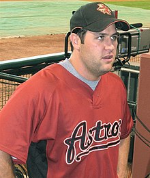 A man in a red baseball jersey with "ASTROS" on the chest and black cap stands in a baseball dugout looking up.
