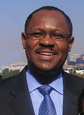 An African-American male with a mustache and glasses wearing a black suit, a blue shirt with white stripes, and a blue tie with silver lines smiles in front of a city skyline.
