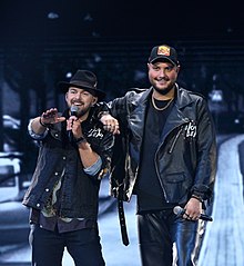 Foto zweier in Schwarz gekleideter Männer, der linke singt in ein Mikrofon