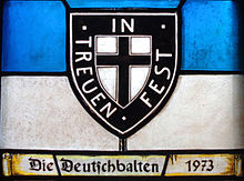 Farbfotografie eines Glasfensters mit blauem und weißem Querstreifen, auf dem ein schwarz-weißes Wappen mit einem schwarzen Kreuz und der Aufschrift „In Treuen fest“ steht. Auf einer ausgerollten Pergamentrolle im unteren Fensterbereich steht „Die Deutschbalten 1973“.
