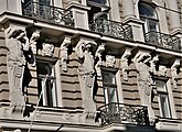 Farbige Nahaufnahme eines Fassadenteils mit drei Fenstern, zwischen denen drei Figuren die Balken der oberen Balkone mit ihren Köpfen und Händen stützen. Ihre Unterkörper bestehen aus Säulen, die mit ihren Oberkörpern verschmelzen.