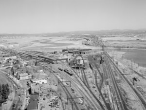 An overview of Cedar Hill Yard, as of 1977. A large collection of railroad tracks can be seen in the foreground. At the center of the image is a large coaling tower, and behind it is a roundhouse. Railroad tracks extend off to the left, as well as off towards the horizon across the Quinnipiac River.