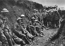 Black and white photo of two dozen men in military uniforms and metal helmets sitting or standing in a muddy trench.