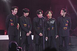 SB19 standing on stage, with each member wearing a black suit while one member giving a speech on a microphone