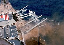 Two large gun turrets are visible, both with guns pointed to the right of the image. The foreground turret is spewing brown colored smoke from the base of all three gun barrels, while water from an unseen hose is being sprayed on the foremost gun barrel.
