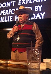 An elderly Caucasian male with a long white mustache. He is wearing a cowboy hat and striped waistcoat while holding a microphone. He is standing in front of a screen.