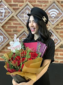 Hòa Minzy wearing a black top, skirt, and hat with Chanel logo, holding a large bouquet of flowers, grinning, turned to right of camera