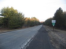 A straight two-lane road between evergreen trees. A signpost at right has signs reading "Cameron County" and "Gibson Township" and "Building-Sewage Permits Required Flood Plain Regulations Enforced" and "10". In the distance is a yellow sign showing a T-intersection at right.