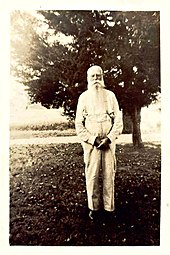 A black and white image (circa 1900s) of an elderly man with white hair and a long beard dressed in a long-sleeved and suspenders standing in front of a tree.