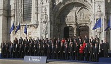 Seitliche Farbfotografie einer Menschengruppe, die in drei Reihen posiert. Fast alle tragen dunkle Anzüge, nur drei Personen haben rote Blazer. Auf einem Schild im Vordergrund steht „Tratado de Lisboa“. Im Hintergrund ist eine gotische Gebäudefassade zu erahnen und mehrere Europaflaggen stehen an beiden Seiten.