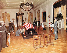 casket draped with a flag in a large room. Several soldiers stand around it, and two clerics kneel on the kneelers at the side. Two empty kneelers are in the foreground