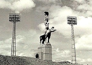 A monumental statue of a nude woman holding a pelvis and riding a tapir, standing between two floodlights.