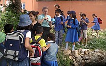 Several children dressed in blue wearing backpacks crowd around a small rock enclosure.