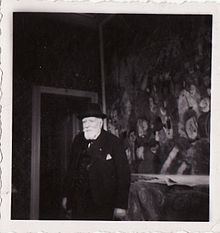 Schwarz-Weiß-Fotografie des alten James Ensor in Anzug, Hut und mit weißem Bart in seinem Haus. An der Wand ist ein Ausschnitt des Gemäldes „Der Einzug Christi in Brüssel“ zu sehen.