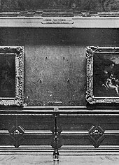 Schwarzweißfotografie einer Ausstellungswand mit zwei alten Gemälden in verzierten Bilderrahmen, die an der rechten und linken Bildseite nur teilweise zu erkennen sind. Zwischen beiden Gemälden ist ein leerer Platz mit vier Nägeln und einem Beschriftungsschild. Im unteren Bereich ist die Wand mit Reliefs verziert.