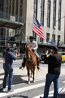 A man in a cowboy hat rides a brown horse. He is carrying a large American flag.