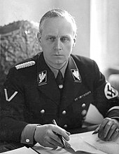 A blonde-haired male wearing a dark Nazi Party uniform with swastika armband seated at a desk