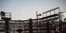 A stadium with three tiers of empty green seats under a pale blue sky, seen through a cast iron fence. At the left a silhouetted man on an elevated platform is at work; at the right, beyond the end of the seating tiers, is a modern high-rise building sheathed in green-tinted glass
