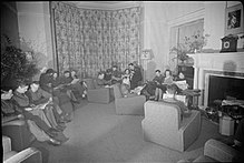 Black and white photograph of men and women sitting on lounge chairs reading newspapers in a large sitting room. Most of the men are wearing military uniform.