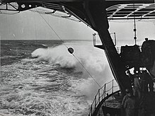Photograph taken on board a ship that shows part of the stern and also the ocean behind the ship. The horizon is in the distance, and there is a large wake behind the ship. The photograph shows a lower deck of the ship as well as the support structure and the bottom of an upper deck. There are about six members of the crew visible at the back of the lower deck who are dwarfed by the ship's structures.