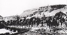 Many fully loaded camels crossing bridge built on square end boats; steep sided mountains in background