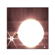 A big round white circle with faint rays around on a brown background. A black irregular shape stands on its left border. A black spot to its left issues six white spikes separated by 60 degrees and two fainter spikes in vertical.