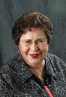 An older white woman, with dark hair, wearing glasses.