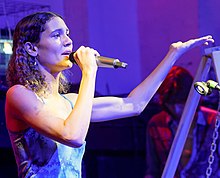 Foto einer Frau, die auf einer Bühne in ein Mikrofon singt und mit ihrer freien Hand gestikuliert. Im Hintergrund ein weiterer Musiker.