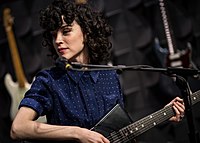 A woman, dressed in blue, looks towards her right and plays a guitar against a black backdrop.
