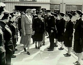 Two women and three men flanked by two lines of women in dark military uniforms