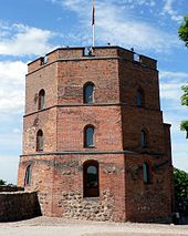 Frontale Farbfotografie eines braunen, dreigeschossigen Turms mit Rundbogenfenstern aus Glas. Teilweise sind größere, ungeformte Steine eingebaut. Auf dem Turm ist eine Aussichtsplattform mit Menschen und einer Flagge.