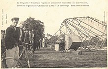 A postcard:On the right, an airship's gondoloa and keel lie on the ground on the side; on the left several people stand looking toward the camera.