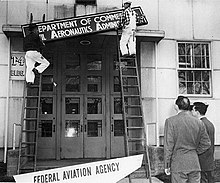 Workmen on ladders are swapping an old "Civil Aeronautics Administration" sign for a new "Federal Aviation Agency" sign above a tall doorway of a building.