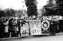 Frontale Schwarzweißfotografie von einer Menschenmenge an einem Bürgersteig. In der Mitte werden drei Plakate mit kyrillischer Schrift „CCCP“ gehalten, was die ehemalige Sowjetunion darstellt. Rechts neben den Plakaten steht ein alter Mann mit Fotoapparat.