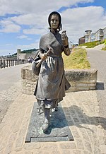 A bronze statue of Mary Anning striding forward, with a pickaxe in one hand and and a fossil in the other.