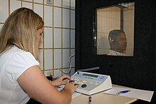 a female medical professional is seated in front of a special sound-proof booth with a glass window, controlling diagnostic test equipment. Inside the booth a middle-aged man can be seen wearing headphones and is looking straight ahead of himself, not at the audiologist, and appears to be concentrating on hearing something