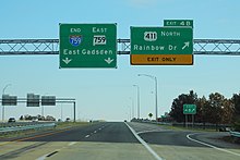Two green signs are located above an elevated portion of roadway with no traffic visible on a cloudy day.
