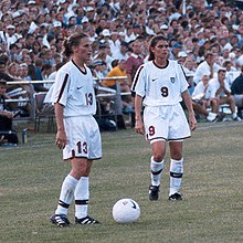 Soccer players Kristine Lilly and Mia Hamm standing over a ball while preparing to take a free kick.