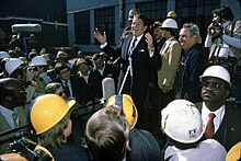 Ronald Reagan stands smiling with his arms outstretched as he speaks to a crowd of men in hard hats.