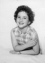A studio pose of a six- or seven-year-old girl with short dark curly hair in a sleeveless print dress.
