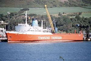 The ship 'Europic Ferry' has an orange-coloured hull and a white superstructure. White lettering names the operator Townsend Thoresen. The ship is moored at a dock next to a verdant hillside.