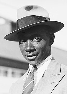 A head and shoulders black and white photograph man in a broad-brimmed hat and suit smiling at the camera.