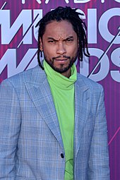 A photo of Miguel, lightly bearded and with thinly braided hair, wearing a blazer over a high-neck sweater