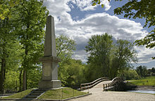 Gray stone obelisk on a rectangular pedestal of the same stone. Behind the monument is a wooden bridge across a river. The Minute Man can be seen in the far background beyond the bridge.